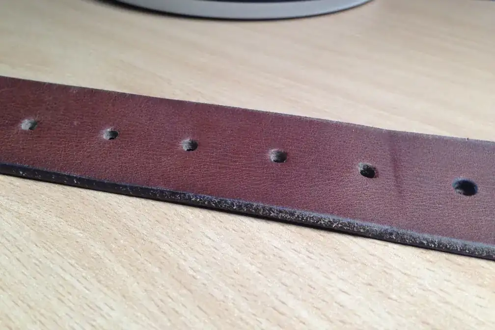 Tramo de un cintur&oacute;n de cuero marr&oacute;n con sus agujeros, apoyado sobre una mesa de madera clara